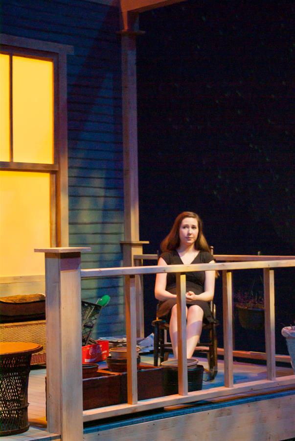 A person sits on a wooden porch at night, illuminated by light from a window. They are in a black outfit, sitting on a chair, with a thoughtful expression. Various objects are around the porch, which has a partially visible house exterior.