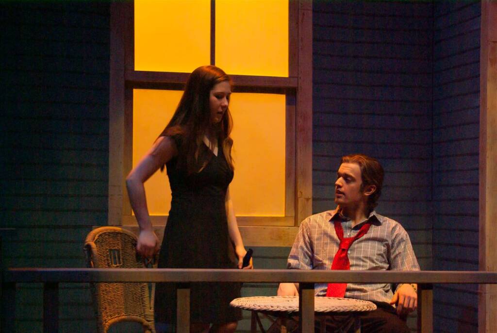 A woman in a black dress stands beside a seated man in a shirt and red tie on a porch. They appear to be engaged in a serious conversation. The background is a dimly lit house with a large window casting a warm glow.