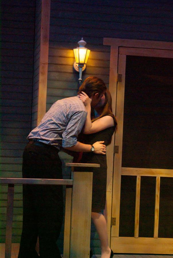 Two people are embracing and kissing on a dimly lit porch at night. A lamp on the wall casts warm light on the scene. The setting suggests a sense of intimacy and romance.