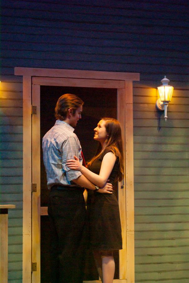 A man and woman stand embraced in front of a wooden door under warm lighting. Both appear to be smiling as they gaze at each other. The surrounding walls are painted in shades of blue, featuring a lit wall lamp on the right.