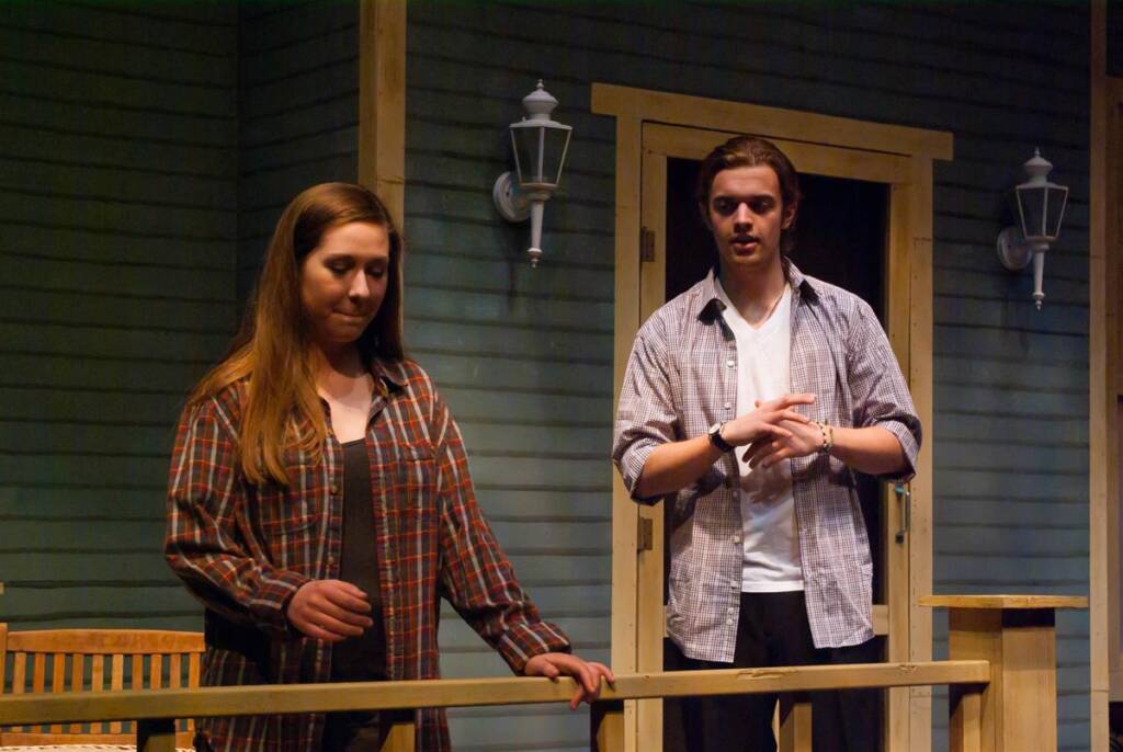 Two people on a wooden porch in front of a house with blue siding. They are wearing casual plaid shirts, and the scene appears to be part of a performance or play. Two wall-mounted lamps flank a door in the background.