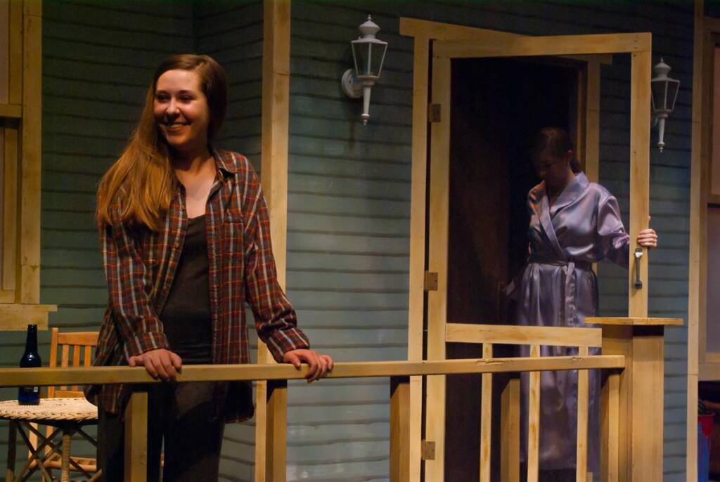 A woman in a plaid shirt smiles while leaning on a wooden porch railing. Another woman in a robe stands in the doorway behind her. The scene is dimly lit, and a bottle is on a nearby chair. The setting has a rustic, relaxed atmosphere.