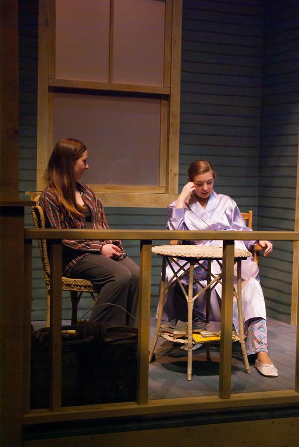 Two women sit on a porch engaged in conversation. One wears a plaid shirt and jeans, the other in a satin robe, resting her chin on her hand. They are surrounded by wooden railing and a window in the background.