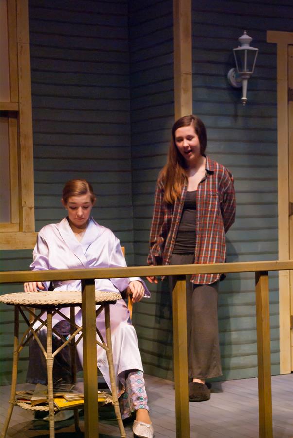 Two people are on a stage set resembling a porch. One is seated at a small table wearing a light robe and patterned pants. The other stands behind, wearing a plaid shirt and dark pants. A wall lamp is mounted on the dark-paneled wall.