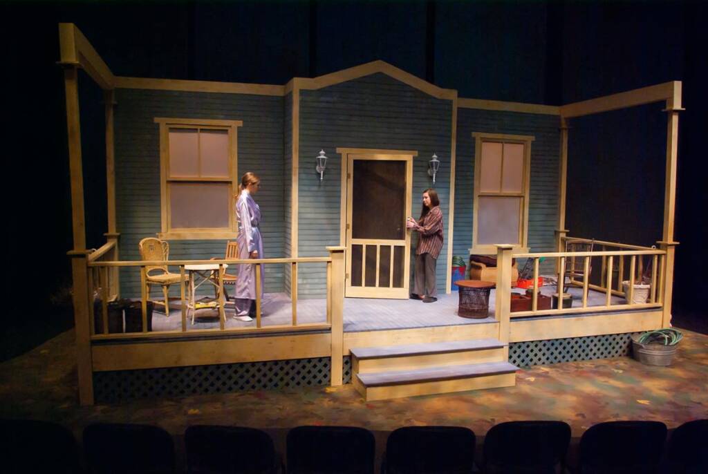 Two people stand on a simple wooden porch set on a stage. One person is in a robe, the other in casual clothes. The porch has two lamps, chairs, and various objects scattered around. The theater seats are visible in the foreground.