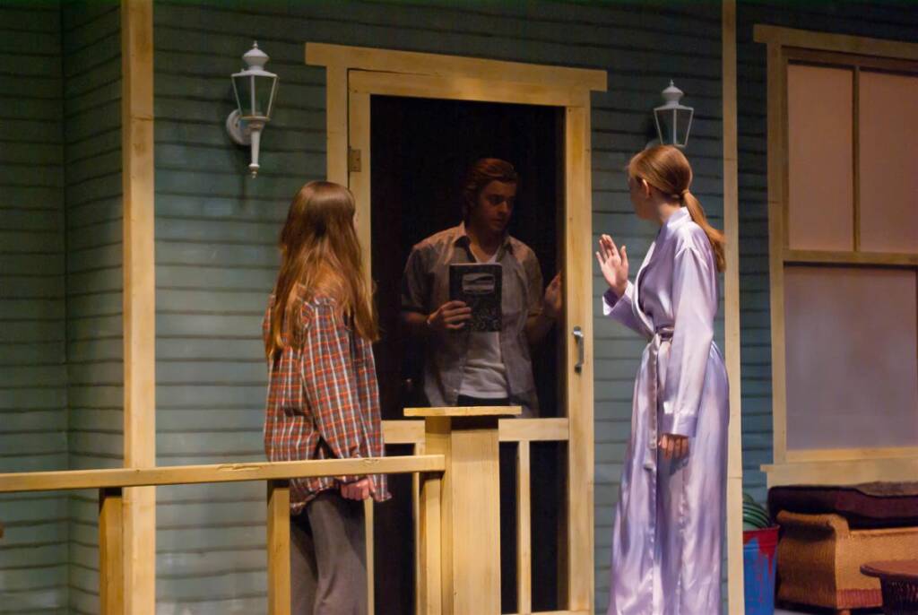 Two women stand on a porch, one in a plaid shirt and the other in a robe. A man is visible through a screen door, holding a book and peering out. The setting is a rustic house with wooden railings and wall-mounted lights.