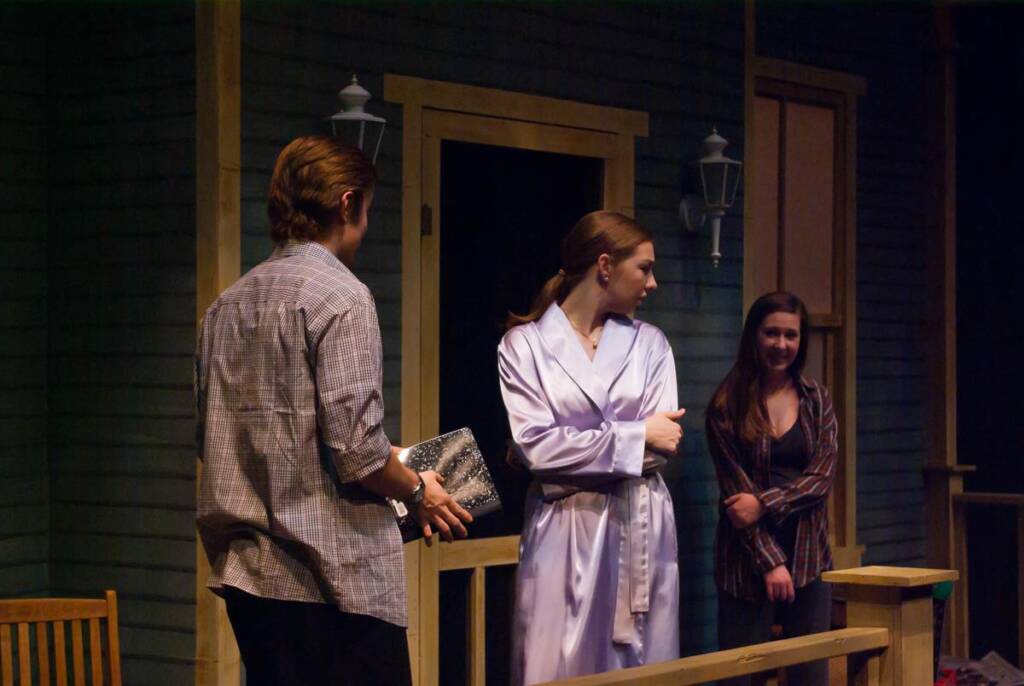 Three people stand on a dimly lit porch. One person, holding a book, and another in a robe, face a third person who is smiling by the door. The setting suggests a casual, possibly dramatic, interaction.