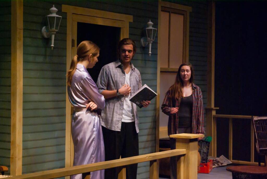 Three people stand on a porch. One wears a light robe, another holds a book, and the third wears a plaid shirt. The setting resembles a stage with a backdrop of a wooden house and decorative lamps.