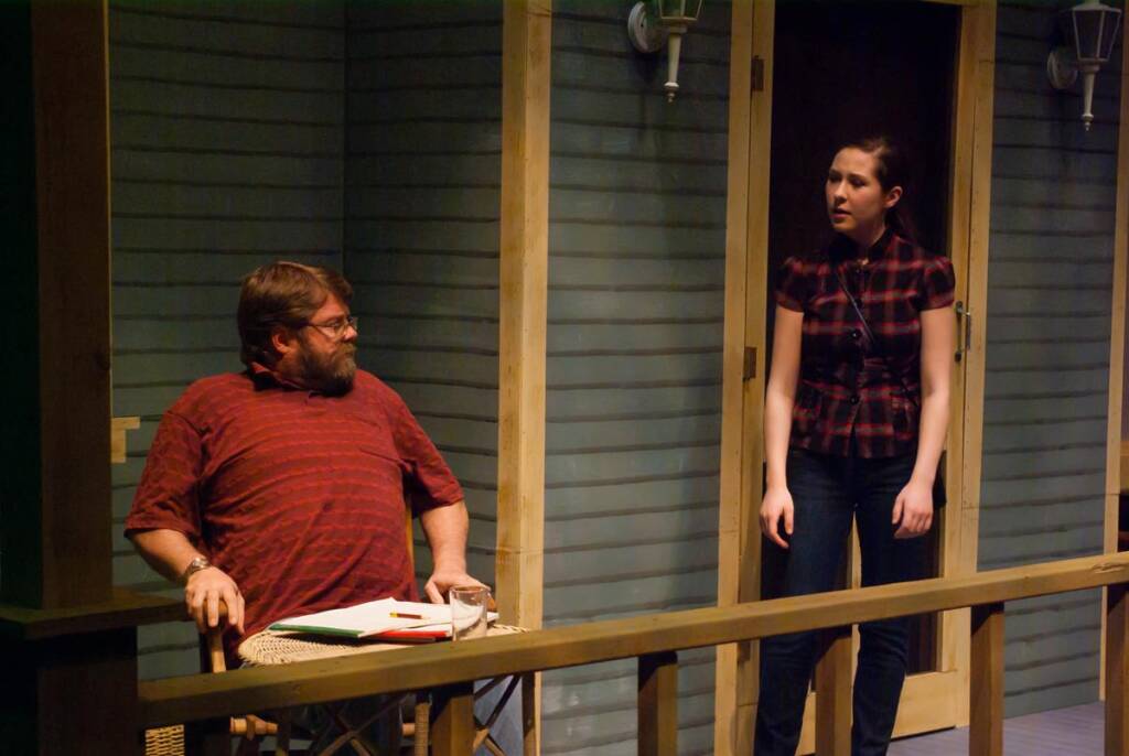 A man with a beard sits at a table on a porch, looking at a woman standing nearby. Both are wearing striped clothing. The porch has a wooden railing and two wall-mounted lamps. There's a notebook and glass on the table. They appear to be conversing.