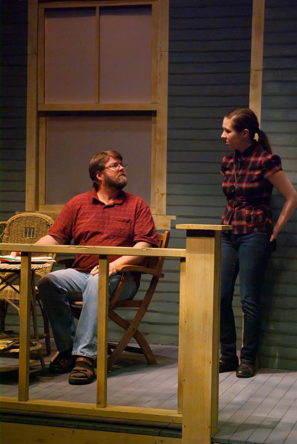 A man with a beard sits on a chair on a porch, looking up at a standing woman. The woman, wearing a plaid shirt and jeans, is leaning slightly forward with her hands behind her back. They are in front of a blue wall with a large window.