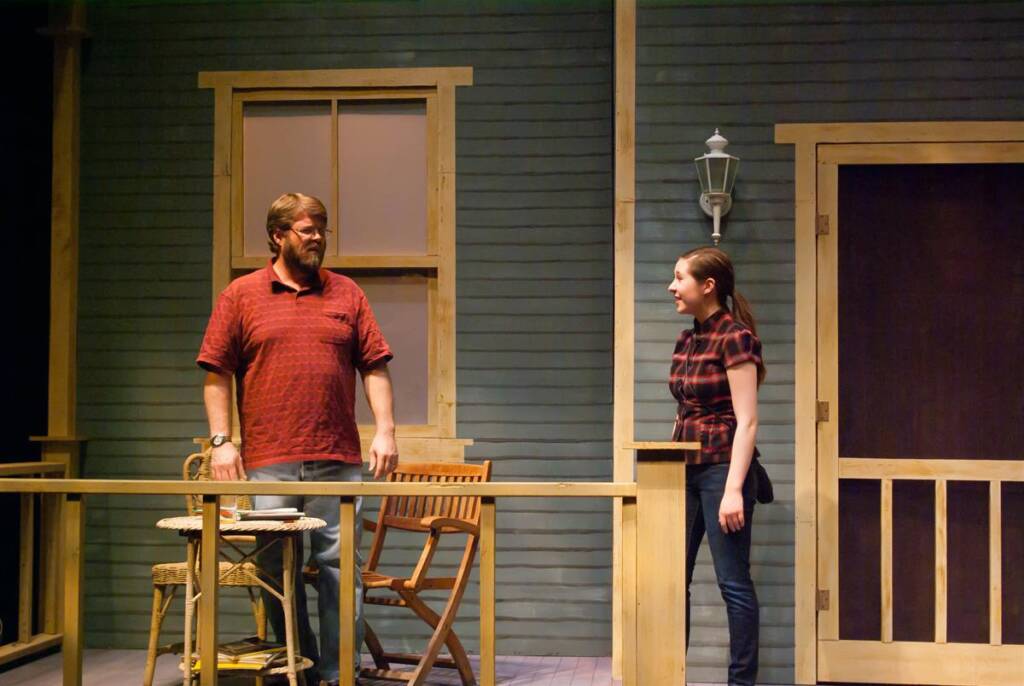 A man in a red shirt and jeans stands on a porch with two wooden chairs and a small table. A woman in a black and red plaid shirt and jeans stands nearby, facing him. Both appear to be in conversation. The porch has a wooden railing, a door, and a window.