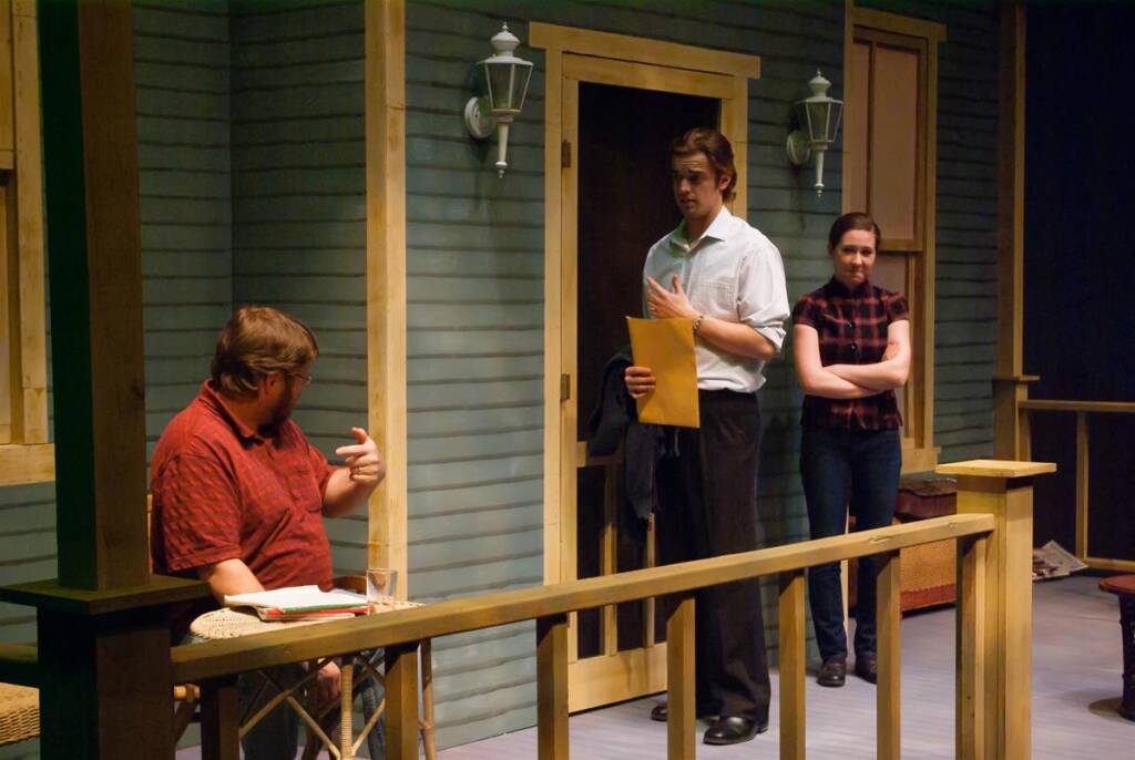 A stage scene depicts three actors. One is seated, pointing, while two others stand on a porch. The man in the middle holds a folder and gestures. The set resembles a wooden house exterior with a door and wall lamps.