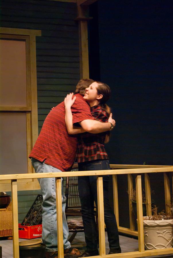 A man and woman embrace in a hug on a porch. The man wears a red shirt and jeans, while the woman wears a plaid shirt and jeans. The setting is a wooden porch with a house wall and potted plant visible in the background.