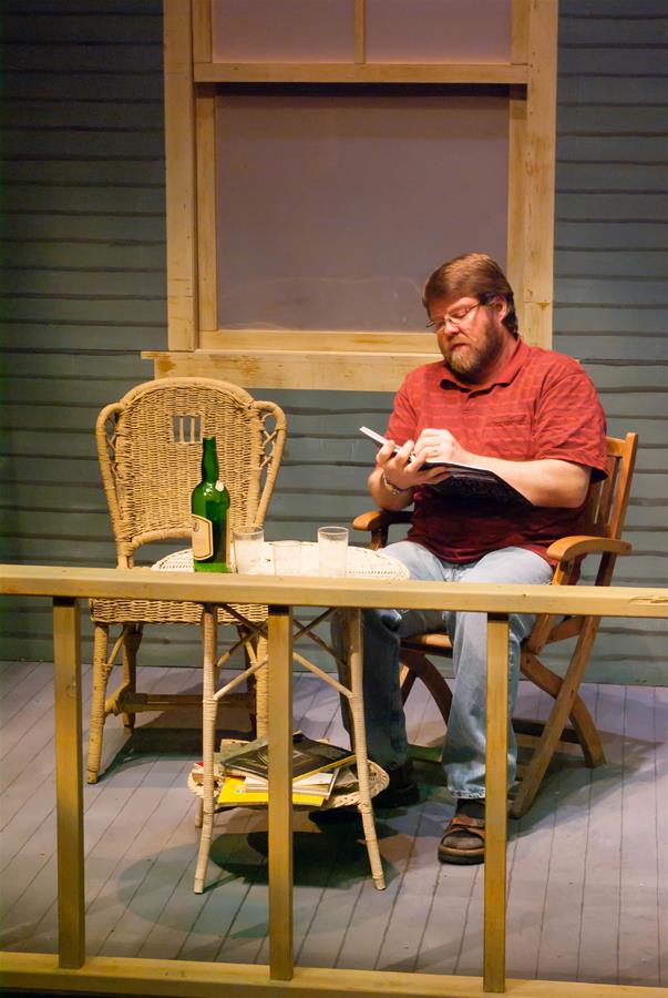 A bearded man in a red shirt sits on a porch, reading a book. Beside him is a small table with a bottle and two glasses. There is a wicker chair and a magazine on the wooden floor. The setting is cozy with a light turquoise wall.