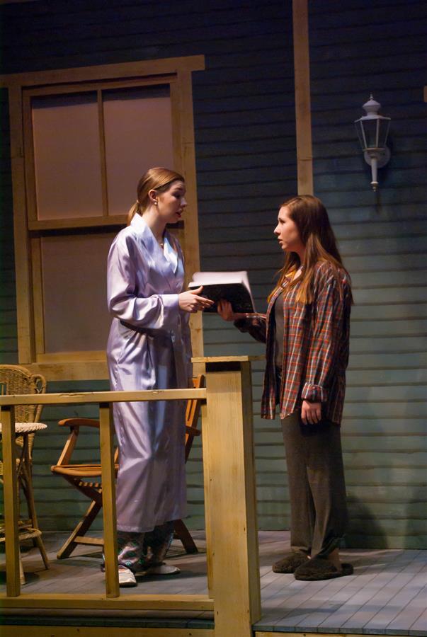 Two women stand on a porch at night. One wears a robe and slippers, holding a book, while the other is in casual clothes and slippers, reaching towards the book. A wooden chair and a wall lamp are visible in the background.