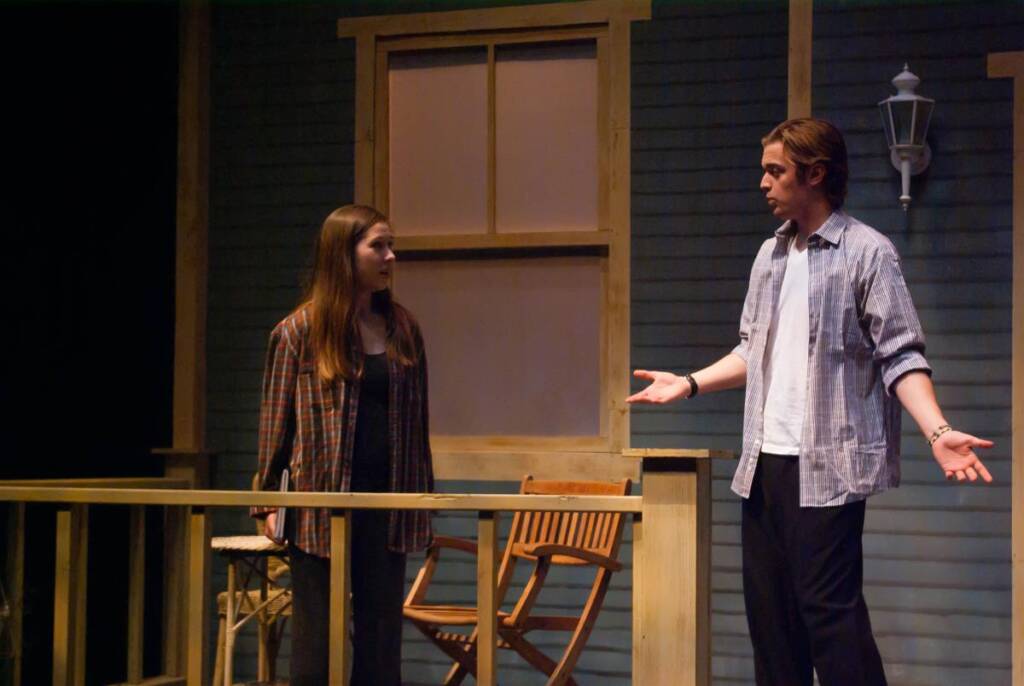 A man and a woman have a conversation on a wooden porch at night. The man gestures with open arms while the woman listens attentively. Both are casually dressed and stand near wooden chairs and a window.