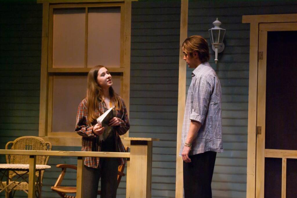 Two people stand on a porch. The woman, holding a notebook, wears a plaid shirt and jeans. The man, in a checkered shirt and dark pants, faces her. A wicker chair and lantern are visible on the house's exterior.