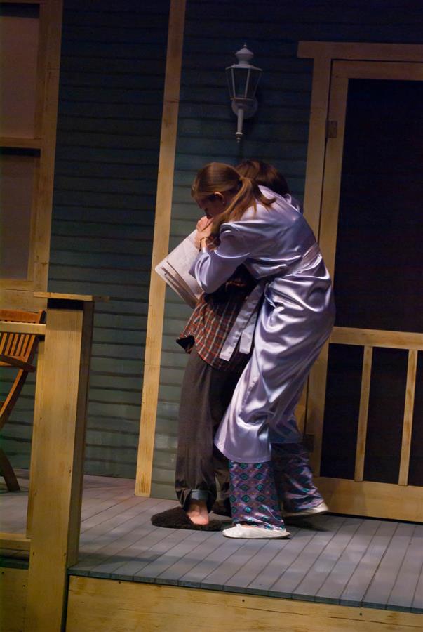 Two people embrace on a porch. One wears a shiny robe and patterned pants, while the other is in dark pajamas. A lamp is mounted on the wall behind them, and there's a wooden railing and chair nearby.