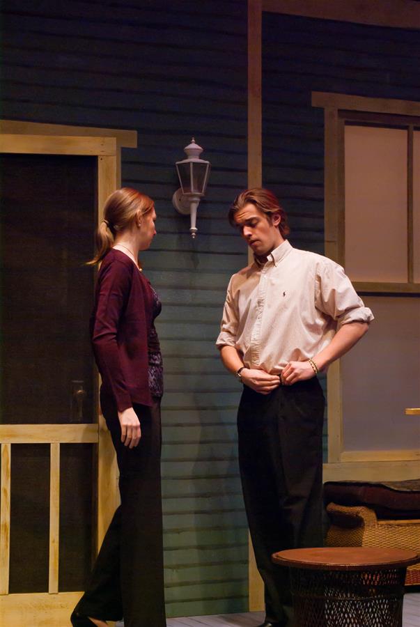 Two actors perform a scene on stage. The woman, with her hair tied back, wears a purple top and dark pants, standing near a wooden door. The man, with short hair, wears a light shirt and dark pants, adjusting his sleeve. The setting resembles a porch.