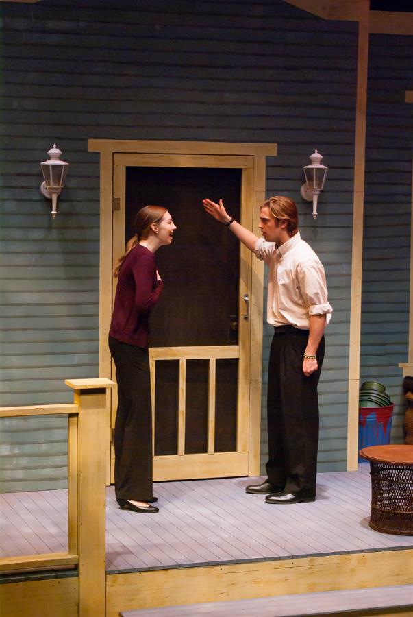 A man and woman are engaged in an intense conversation on a porch with a closed door behind them. The man gestures emphatically with one hand raised, while the woman looks at him, appearing focused. Lanterns are mounted on the wall beside the door.