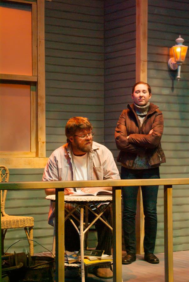 A bearded man sits at a small table on a porch, reading a newspaper. A woman stands beside him, arms crossed, wearing a coat. The porch is warmly lit, with a wicker chair and a wall-mounted light in the background.