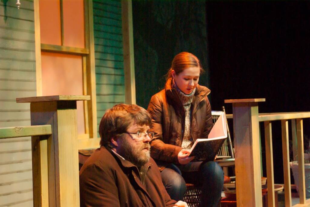 A man with glasses and a beard sits on the steps of a porch, looking thoughtful. A woman in a brown jacket and scarf sits beside him, reading from an open book. The background shows a wooden house exterior.