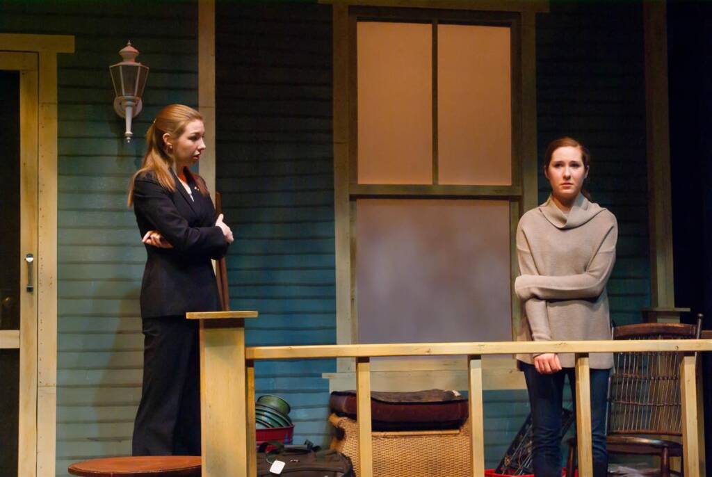 Two women stand on a wooden porch during a stage play. One woman in a suit stands with arms crossed, while the other, in a sweater, leans lightly on the railing. The setting includes bags and a wicker basket, with a closed door to the left.