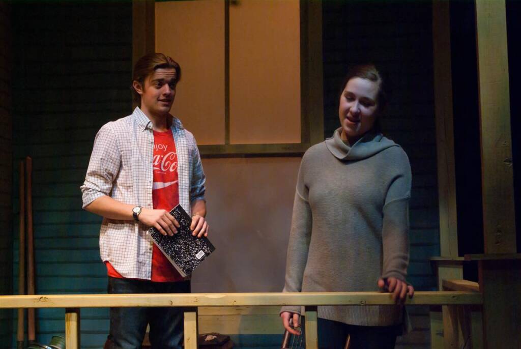 Two people stand on a wooden porch. The person on the left, in a checkered shirt and red T-shirt, holds a notebook. The person on the right, wearing a gray sweater, leans on the railing. The scene appears to be part of a stage performance.