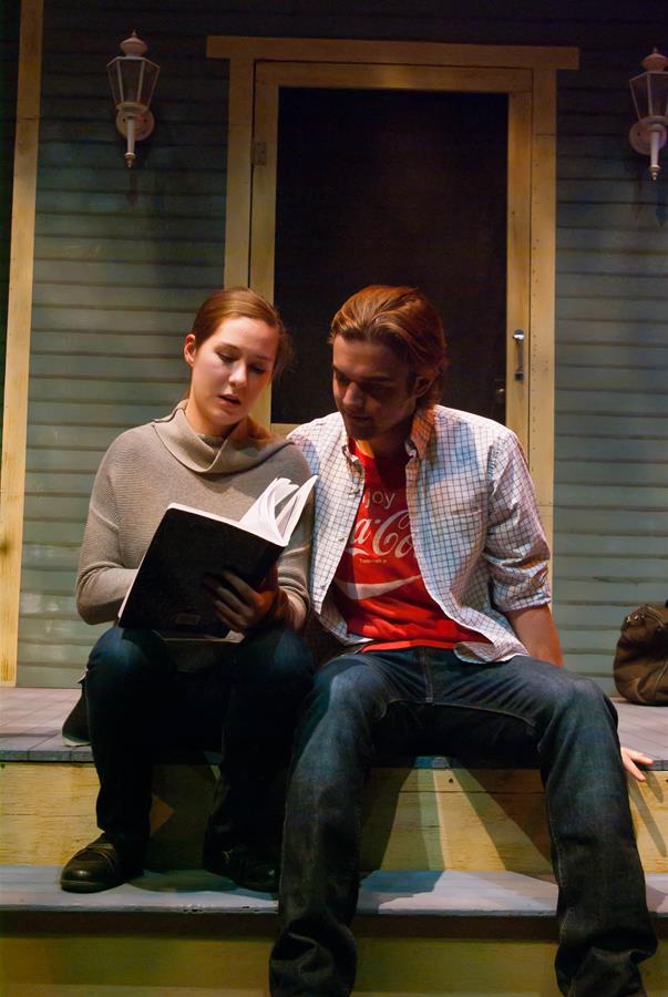 Two people sitting on steps in front of a door. The person on the left, wearing a sweater, is holding an open book. The person on the right, wearing a plaid shirt over a red T-shirt, is looking at the book. Two wall lamps are above them.