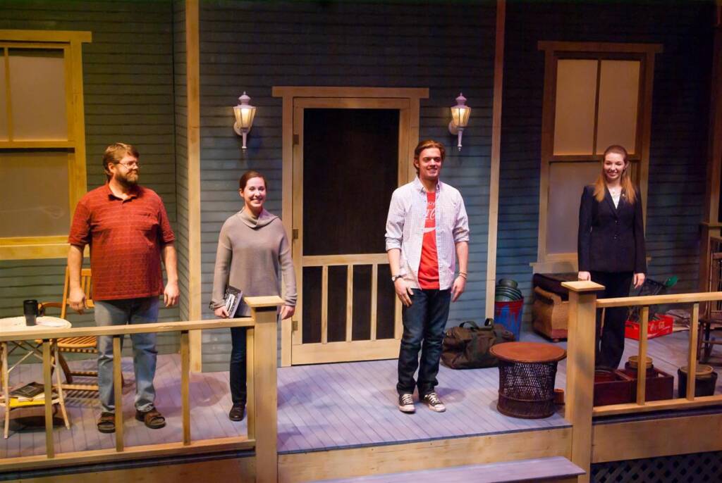 Four people stand on a wooden porch set in a theater. They are positioned along the porch railings, smiling. The set includes a door, windows, a bench with props, and light fixtures. The background suggests a rustic, homey setting.