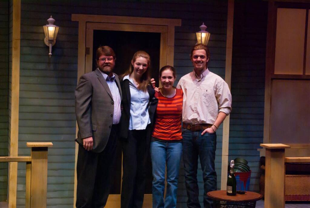 Four people are smiling and posing in front of a house set with blue walls and two wall lamps. They stand closely together, with one person in a suit and others in casual attire. A small table with a champagne bottle and bucket is beside them.