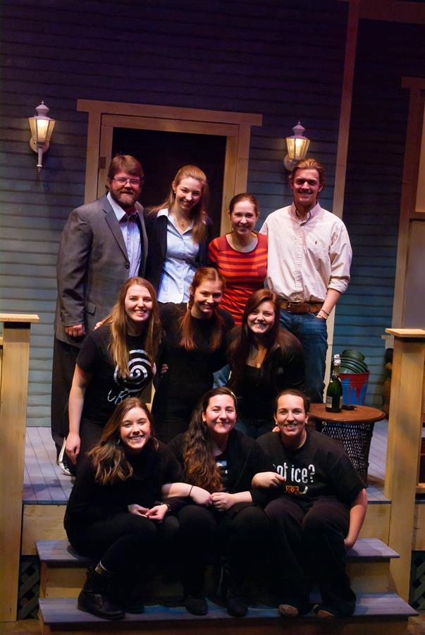 A group of ten people, casually dressed, pose on the steps of a porch. The backdrop resembles a house with two lit wall lamps. Some individuals are standing, while others are sitting, all smiling at the camera.