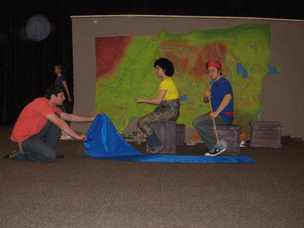 Three people are performing a theater play. One person is kneeling on the left holding a blue fabric, while the other two sit on crates with one holding a stick. A colorful map-like backdrop is behind them. A person walks in the background.