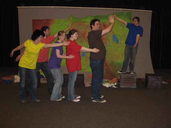 Six people are lined up, each with arms on the shoulders of the person in front. They're striking playful poses against a colorful backdrop. One person stands on a box with arms raised, leading the group. Casual attire suggests a fun, informal setting.