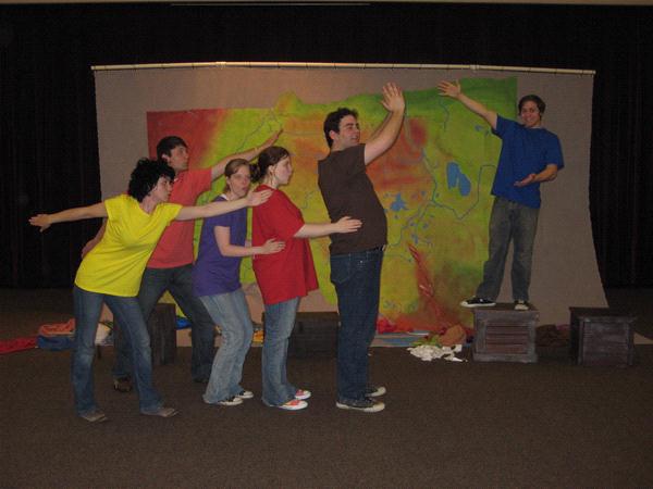 Six people in colorful shirts perform a playful, theatrical pose in front of a colorful map backdrop. Five stand in a line mimicking a swaying motion, while one on the right, in a blue shirt, reaches out with both arms.