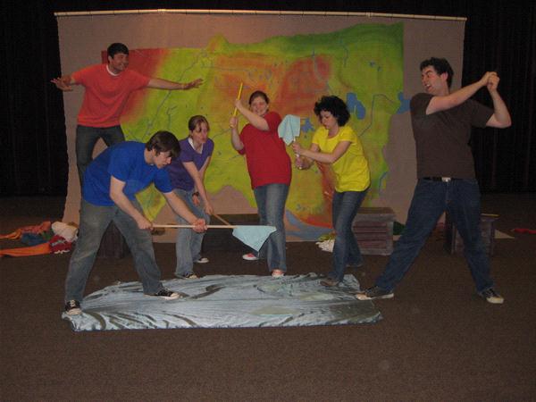 Six people are playfully posing in front of a colorful backdrop. They are standing on a blue sheet on the floor, holding objects like sticks and cloth, with some pretending to row or steer. The backdrop features abstract, bright colors.