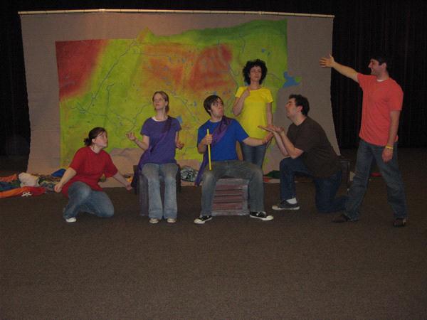 A group of six people perform a skit in front of a colorful backdrop. They wear bright shirts and are in animated poses, some kneeling or standing, gesturing expressively. The backdrop features abstract, multicolored designs.