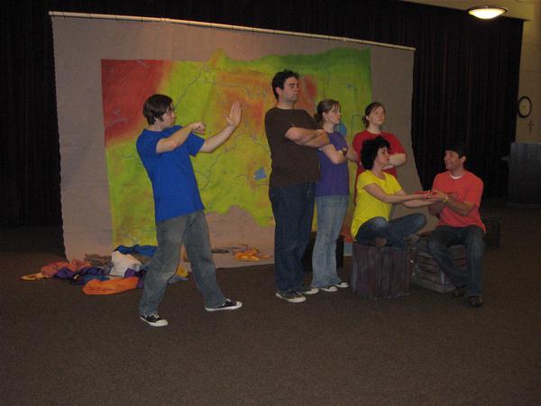 Five people perform a skit in front of a colorful backdrop. One person in a blue shirt gestures forward, while two in the center stand with folded arms. Two others sit on the right, engaged in conversation, surrounded by colorful props.