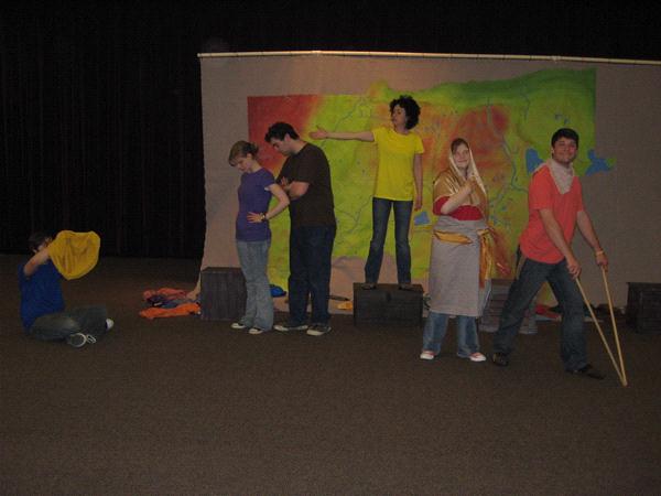 A group of people perform a play on stage. One person sits to the left with a cloth, while others stand forming various poses. The background features a colorful mural. The setting appears informal, with casual clothing and minimal props.