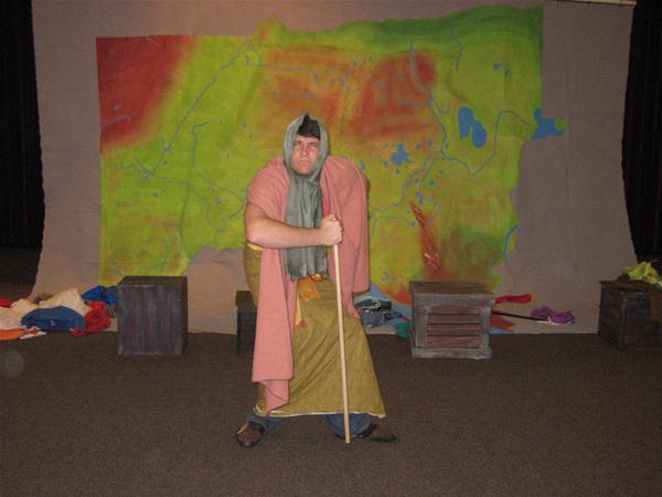 A person dressed in layered, colorful robes poses with a wooden staff in front of a large map backdrop. Crates and fabrics are scattered on the floor. The person has a humorous expression, enhancing a theatrical or playful setting.