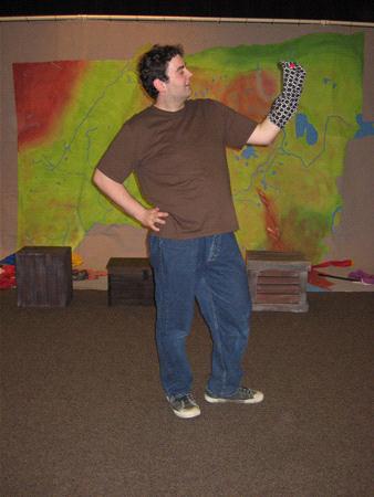 A person wearing a brown shirt and blue jeans poses with a patterned sock on their hand, against a colorful abstract backdrop. Three boxes are on the floor behind them.