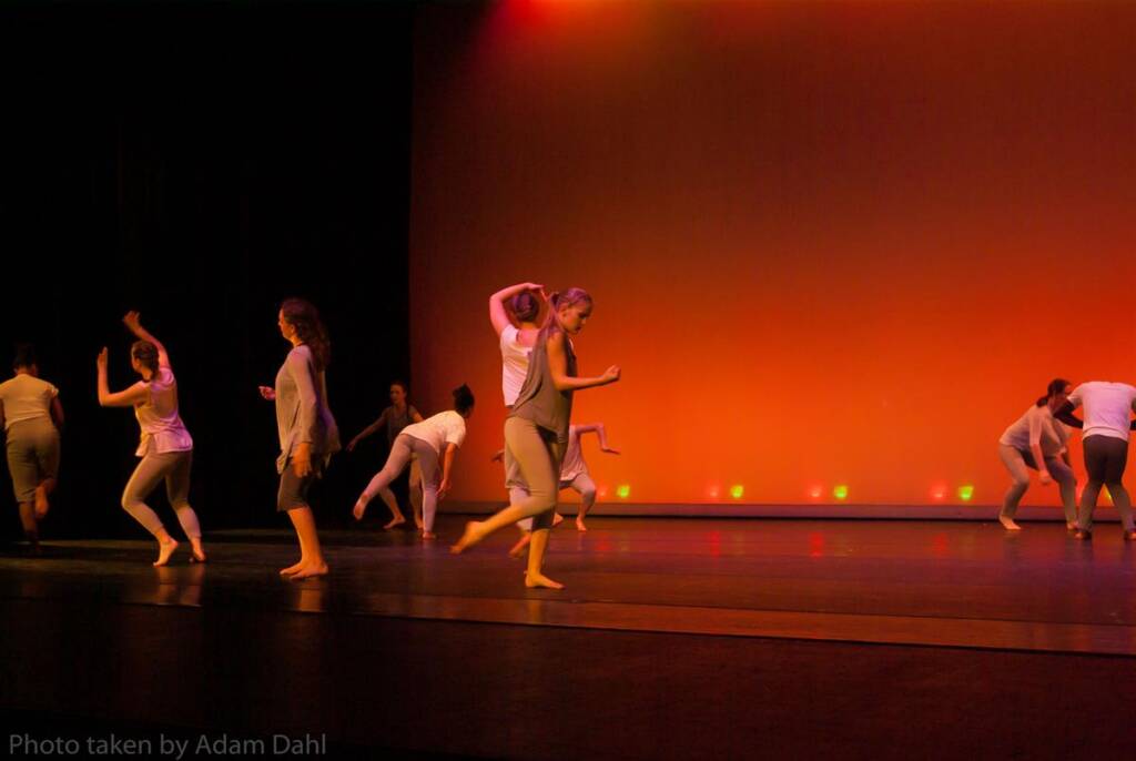 Dancers in casual attire perform on stage against an orange-lit backdrop. Their movements are dynamic and fluid, creating an expressive contemporary dance scene. The stage lighting casts soft shadows, enhancing the vibrant atmosphere.