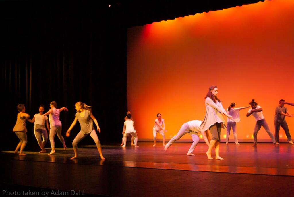 Dancers in casual clothing perform a contemporary routine on a stage with warm orange lighting. Some are mid-motion while others hold still poses, creating a dynamic and varied composition.