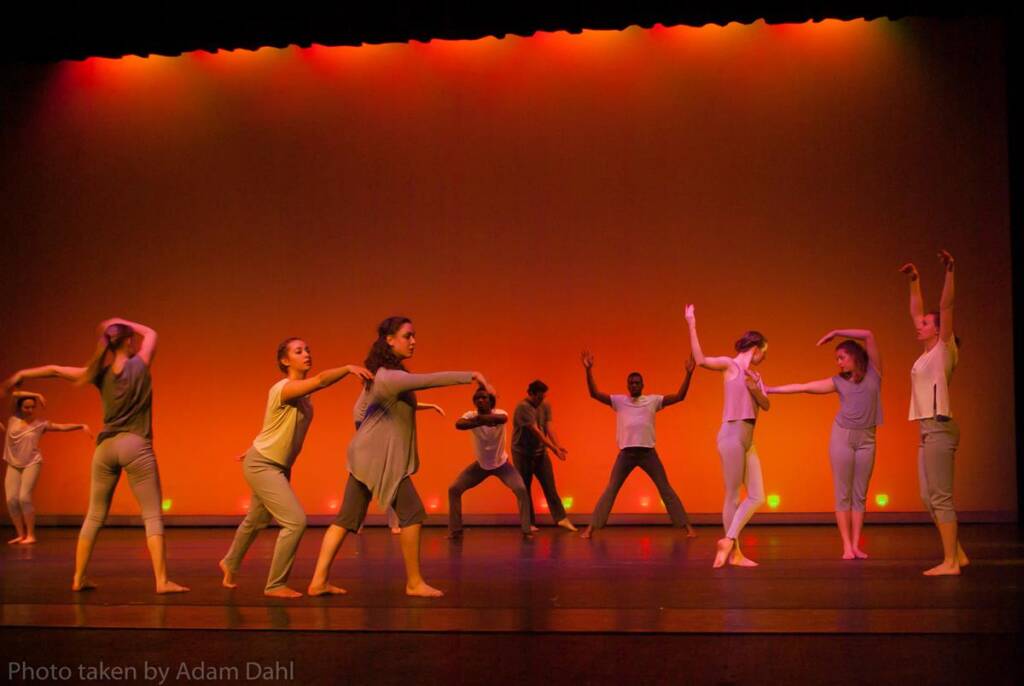 A group of dancers in casual outfits perform a contemporary dance on stage, set against a warm orange backdrop. They strike dynamic poses, extending arms and legs, creating a sense of movement and energy. The lighting highlights their expressive gestures.