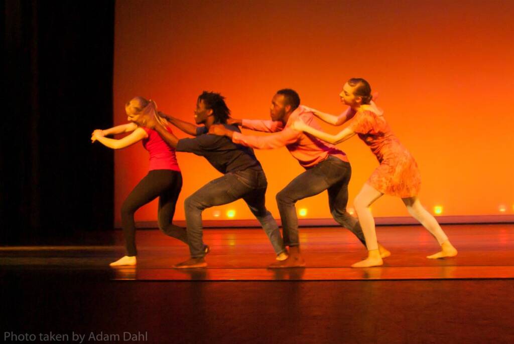 Four dancers perform on stage against an orange backdrop. They form a line, each holding the waist of the person in front. The dancers wear casual clothing in various colors, and the scene is warmly lit.