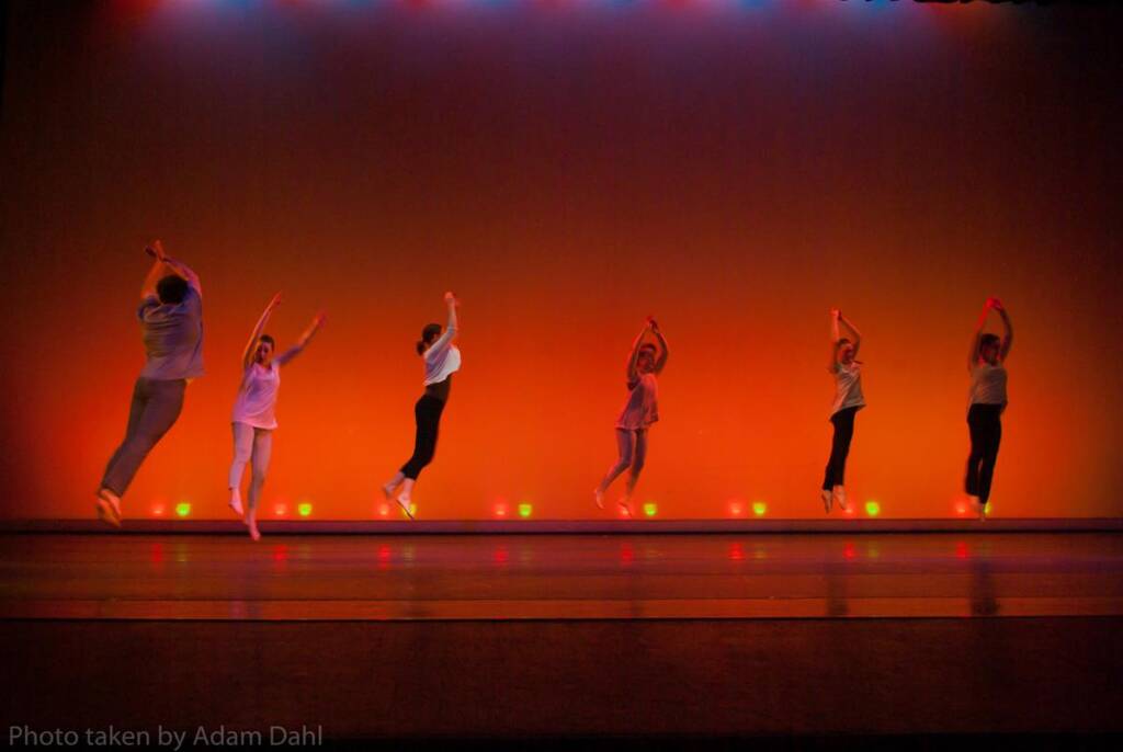 Six dancers leap in unison on stage, silhouetted against a warm orange background with colorful lights above. Their movements are synchronized, creating a sense of energy and rhythm.