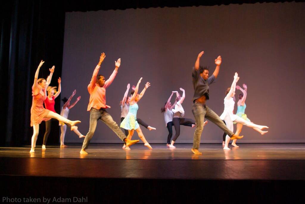 A group of diverse dancers perform energetically on stage, each with one leg lifted and arms raised. They wear colorful outfits, adding vibrancy to the scene. The background is a plain, dark backdrop, emphasizing their movements.