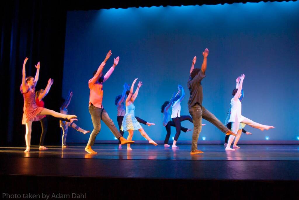 Dancers on stage perform in dynamic poses with arms extended and legs lifted. They wear colorful costumes and are illuminated by blue stage lighting, creating a vibrant and energetic scene.