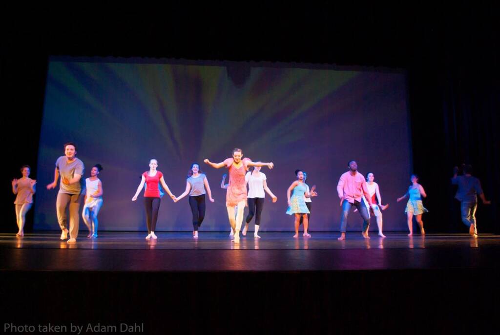 A group of dancers performs on stage, wearing bright and colorful outfits. They are mid-motion, and the background is lit with blue and yellow lights, creating a dynamic and lively atmosphere.