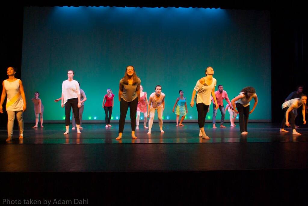 A group of dancers, wearing a mix of colorful and neutral outfits, perform energetically on a stage with a green backdrop. They are in various poses, suggesting movement and rhythm.